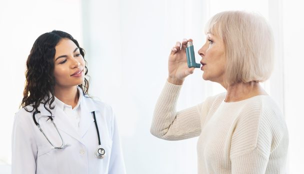 Medical Advice. Elderly woman using asthma inhaler, mexican doctor watching, free space, panorama